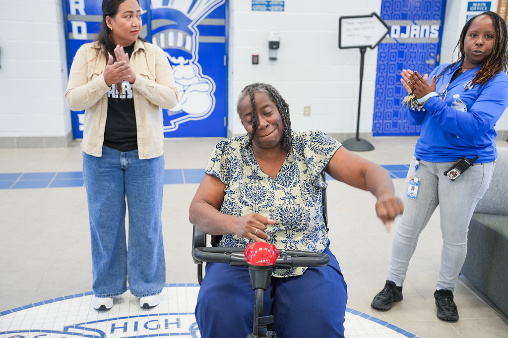 A woman sits on a scooter in a hallway with blue walls, while two women clap behind her.