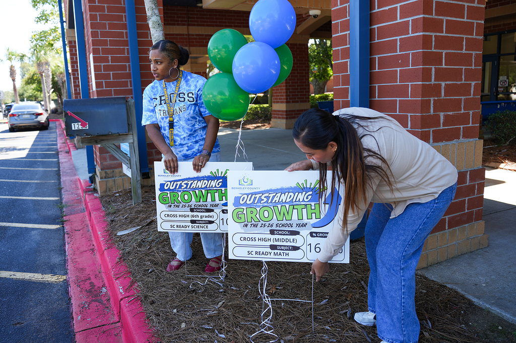 Two women planting a sign near a building. One woman holds a sign that says "Outstanding Growth."