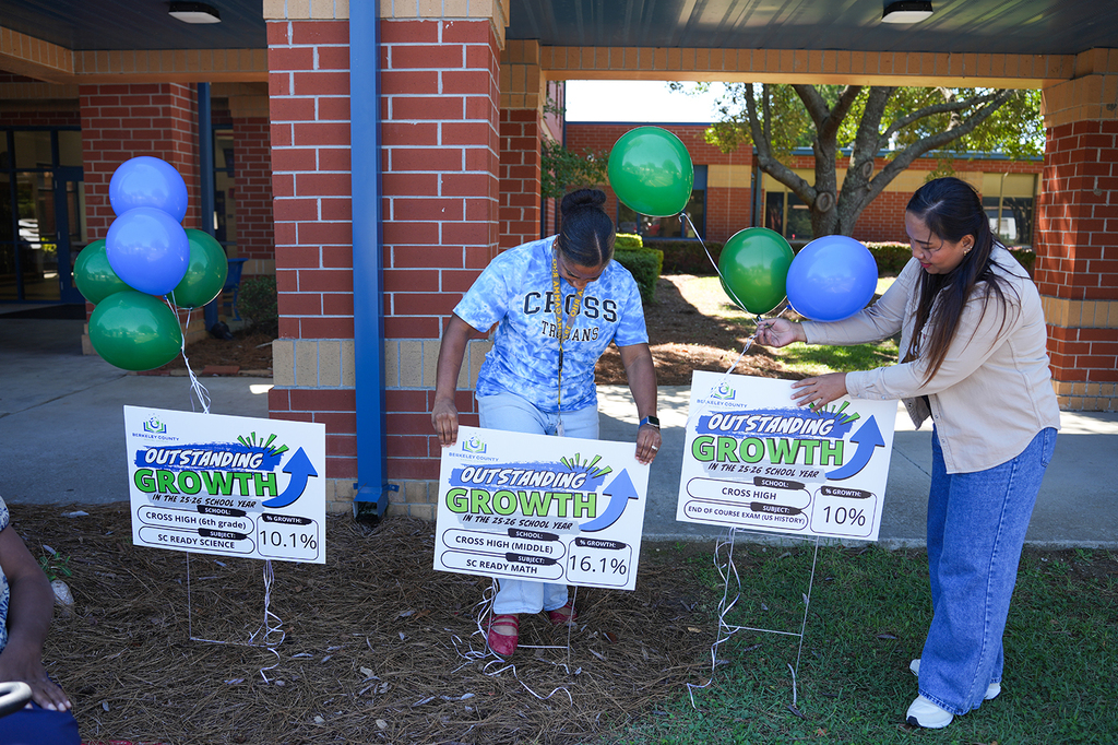 Two people hold signs reading "Outstanding Growth" in front of a brick building. Each holds balloons.