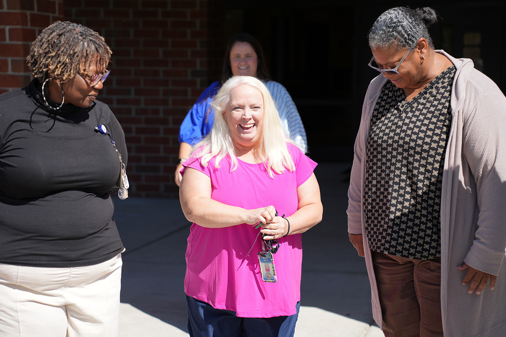 Four women outside a building, one holds keys, two wear glasses, one in pink, two in black tops.