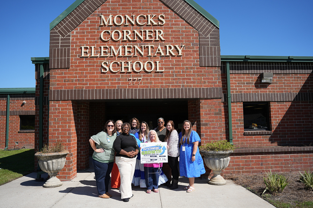 Eleven adults and one child stand in front of Moncks Corner Elementary School. They hold a large banner.
