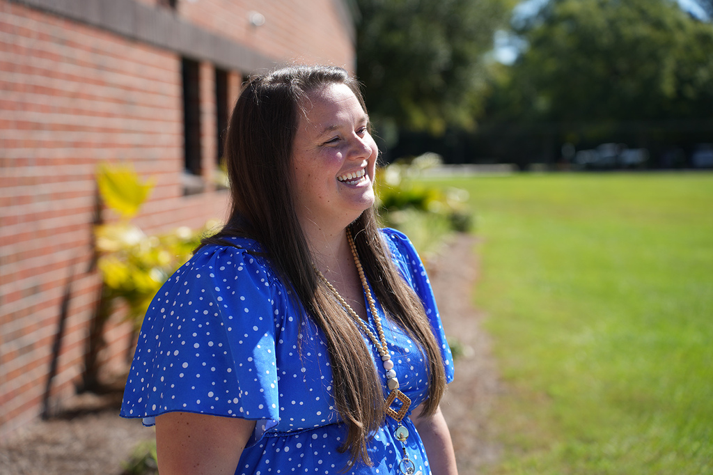A woman stands by a brick building, smiling. Behind her is a lawn and greenery. The sun casts shadows on the wall.
