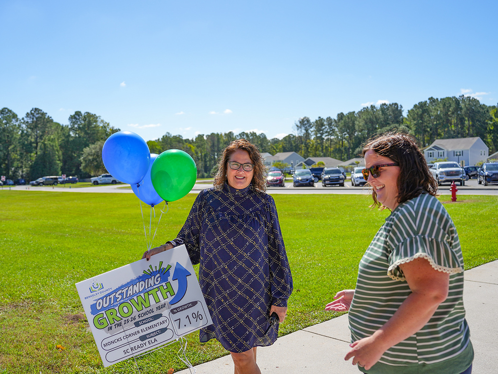 Two women on a sidewalk with a grassy field in the background. One woman holds a banner and colorful balloons.