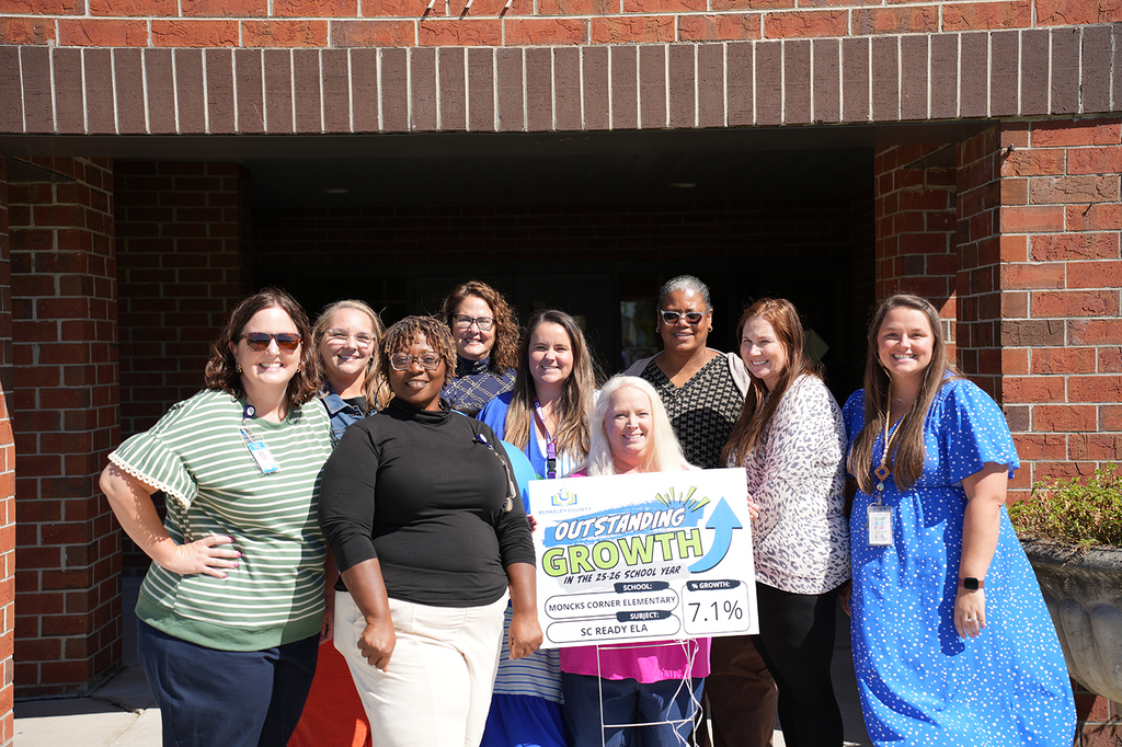 A group of eight people poses in front of a brick building, holding a sign with an arrow pointing upward.