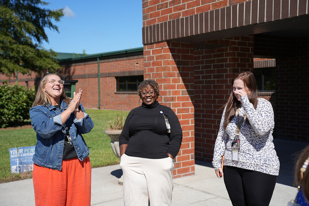 Three women stand outdoors, one clapping. The building has a brick wall and glass windows. Green grass and a potted plant are in the background.