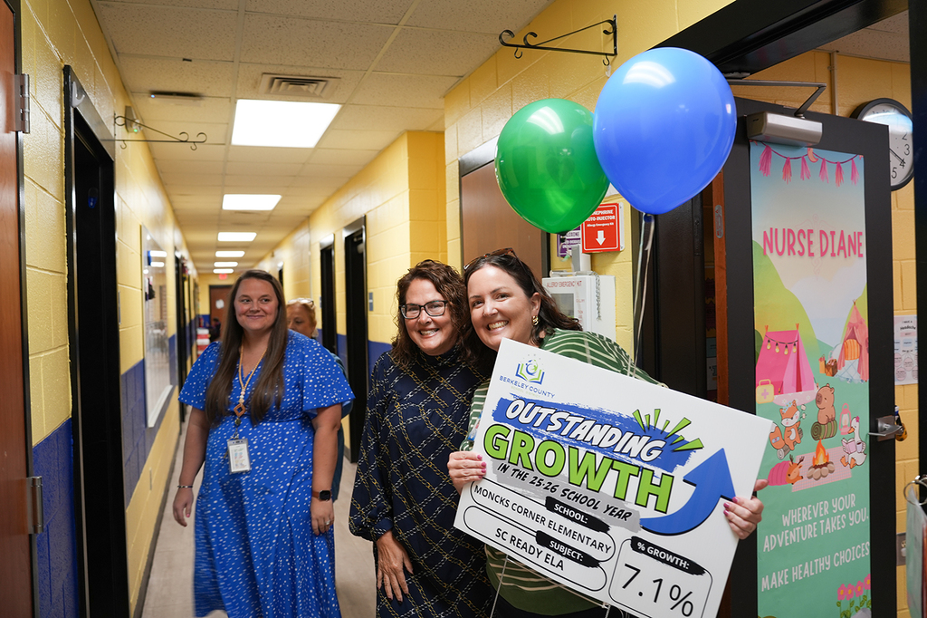 Three women in a hallway hold a sign. One holds a banner, another holds balloons. Background has yellow and white walls and a door.