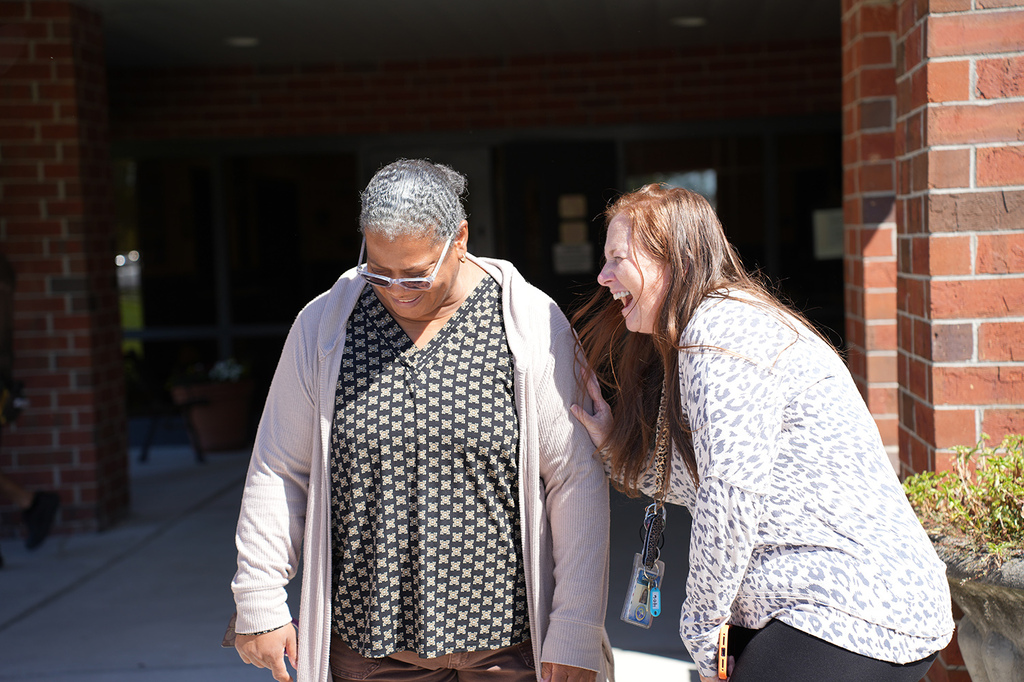 Two women laughing outside a building with brick walls. One wears glasses, a polka-dot top, and a cardigan.