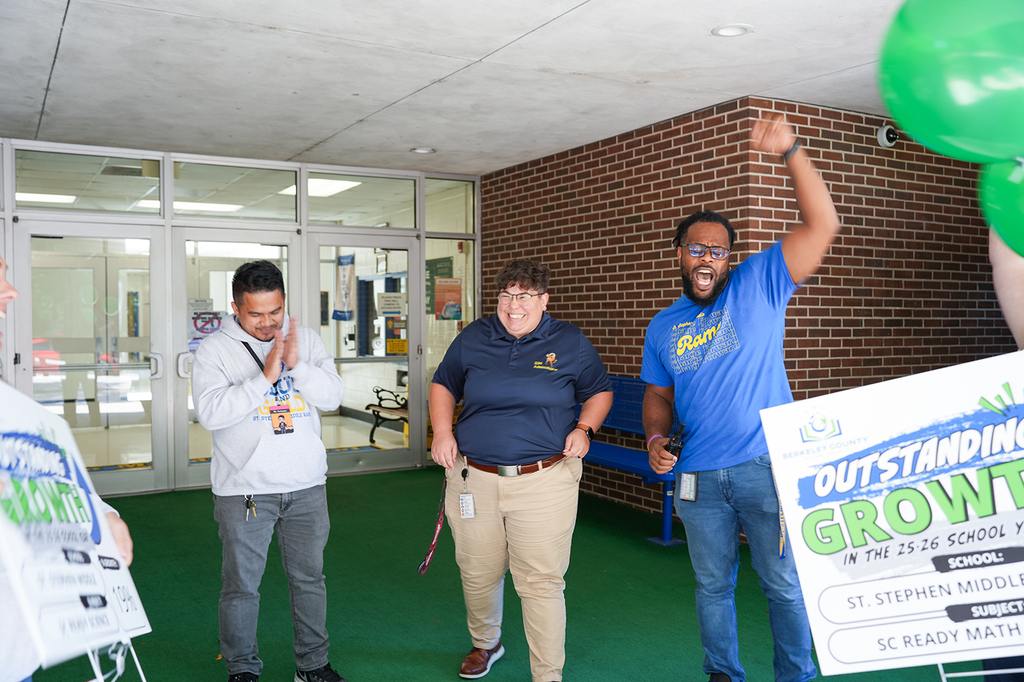 Four people standing in a room with green balloons and signs. A brick wall and glass doors in the background.