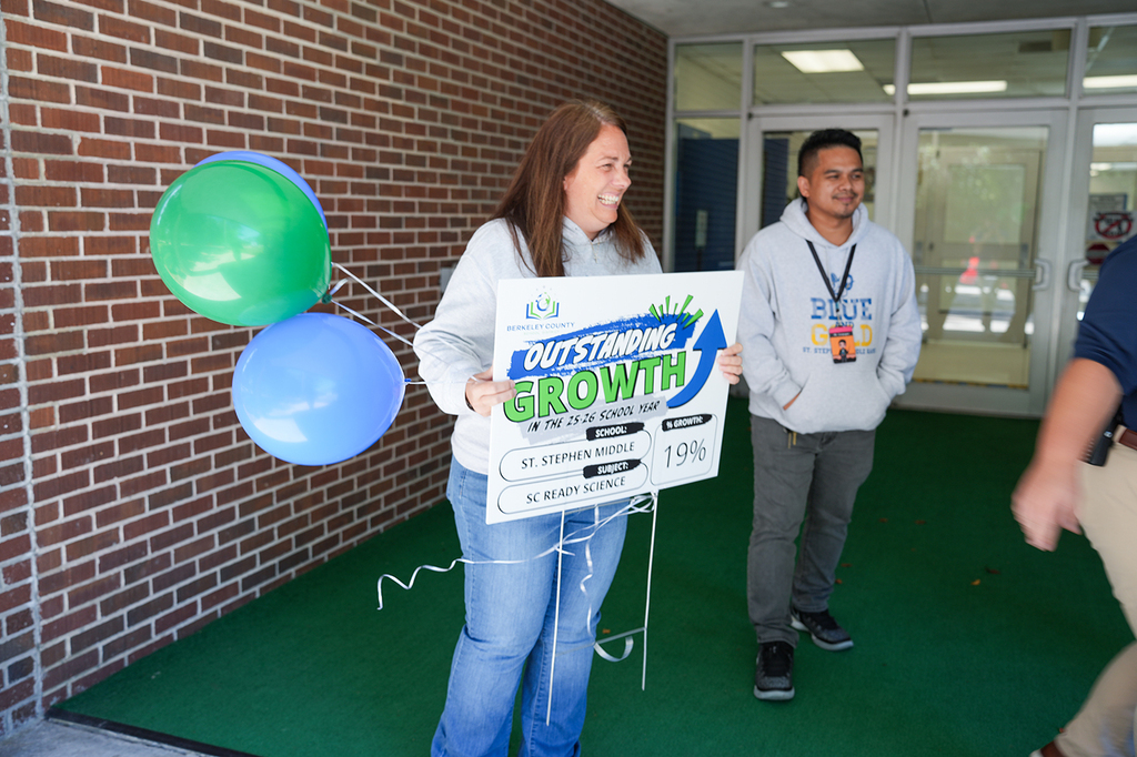 A woman holds a sign saying "Outstanding Growth" with two blue and green balloons. A man stands behind her.