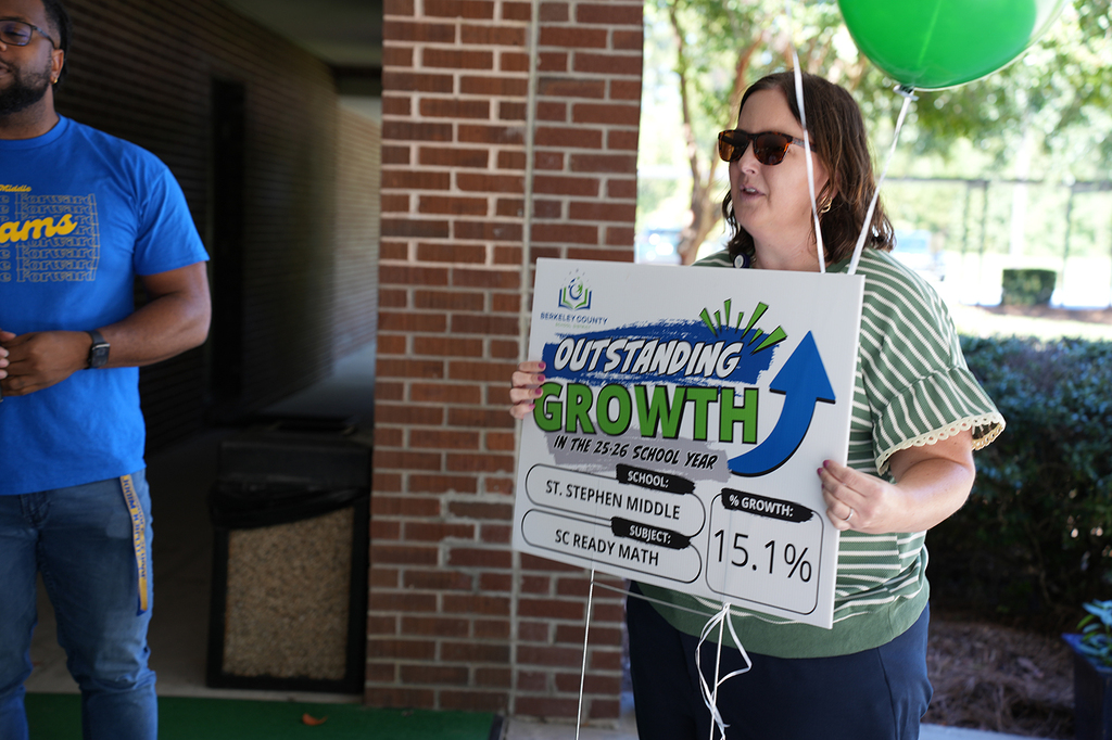 A woman in a striped shirt holds a board with "OUTSTANDING GROWTH" and "15.1%" while standing next to a man.