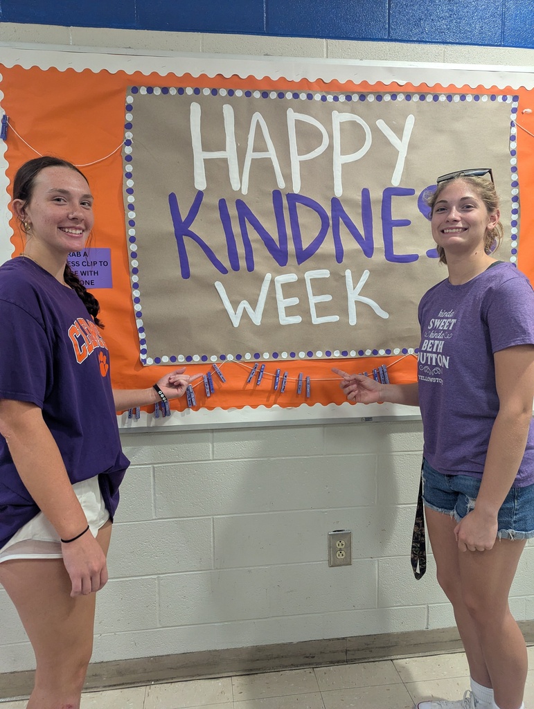 Two young women in purple t-shirts stand next to a bulletin board with the words "Happy Kindness Week."