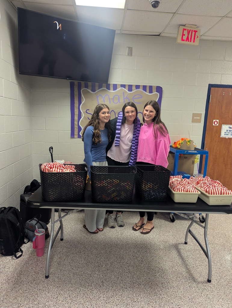 Four people stand behind a table with food. A banner and TV are on the wall.