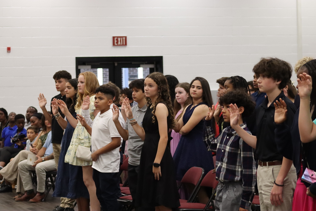 A group of children and adults stand with hands over hearts. Some wear dresses, shirts, and pants.