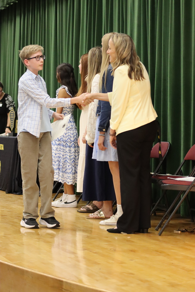 A young boy shakes hands with an adult woman on a stage. Behind them, several girls stand, and green curtains and chairs are visible.