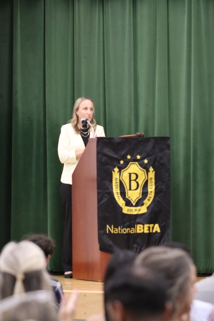Mrs. Johnson speaks at podium with National Beta Club banner. A group of people are listening.
