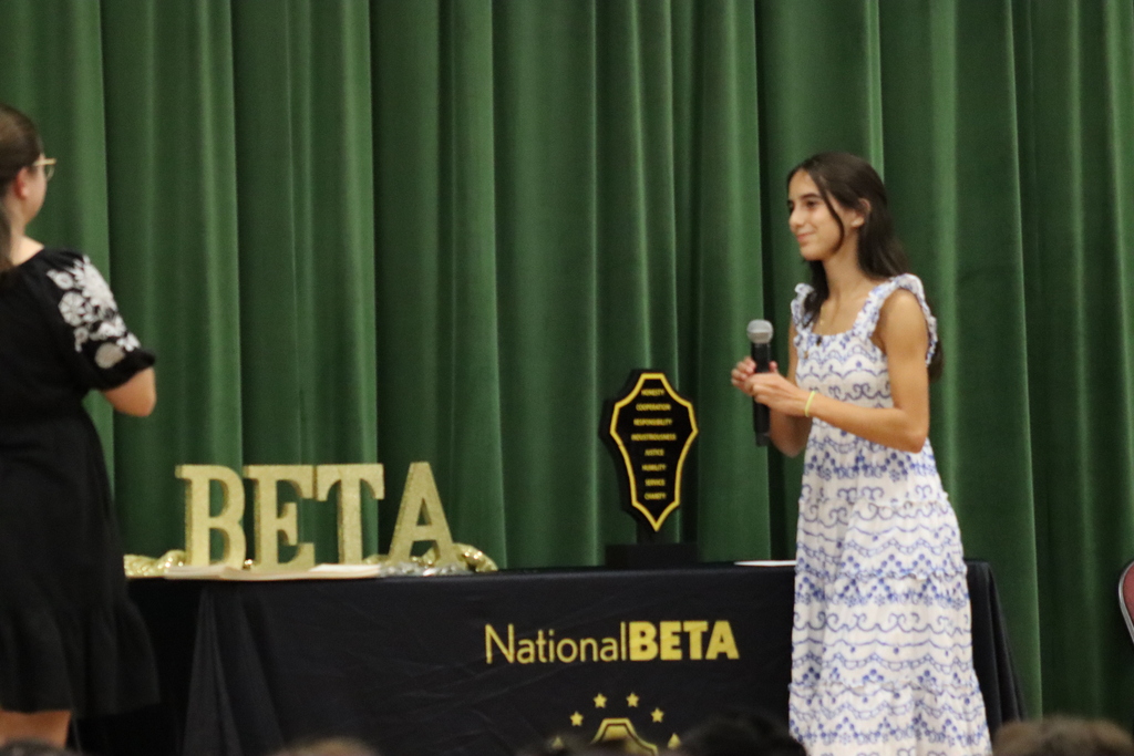 A woman in a dress holds a microphone and speaks on stage. Another woman stands beside a table with a National BETA sign and a trophy.