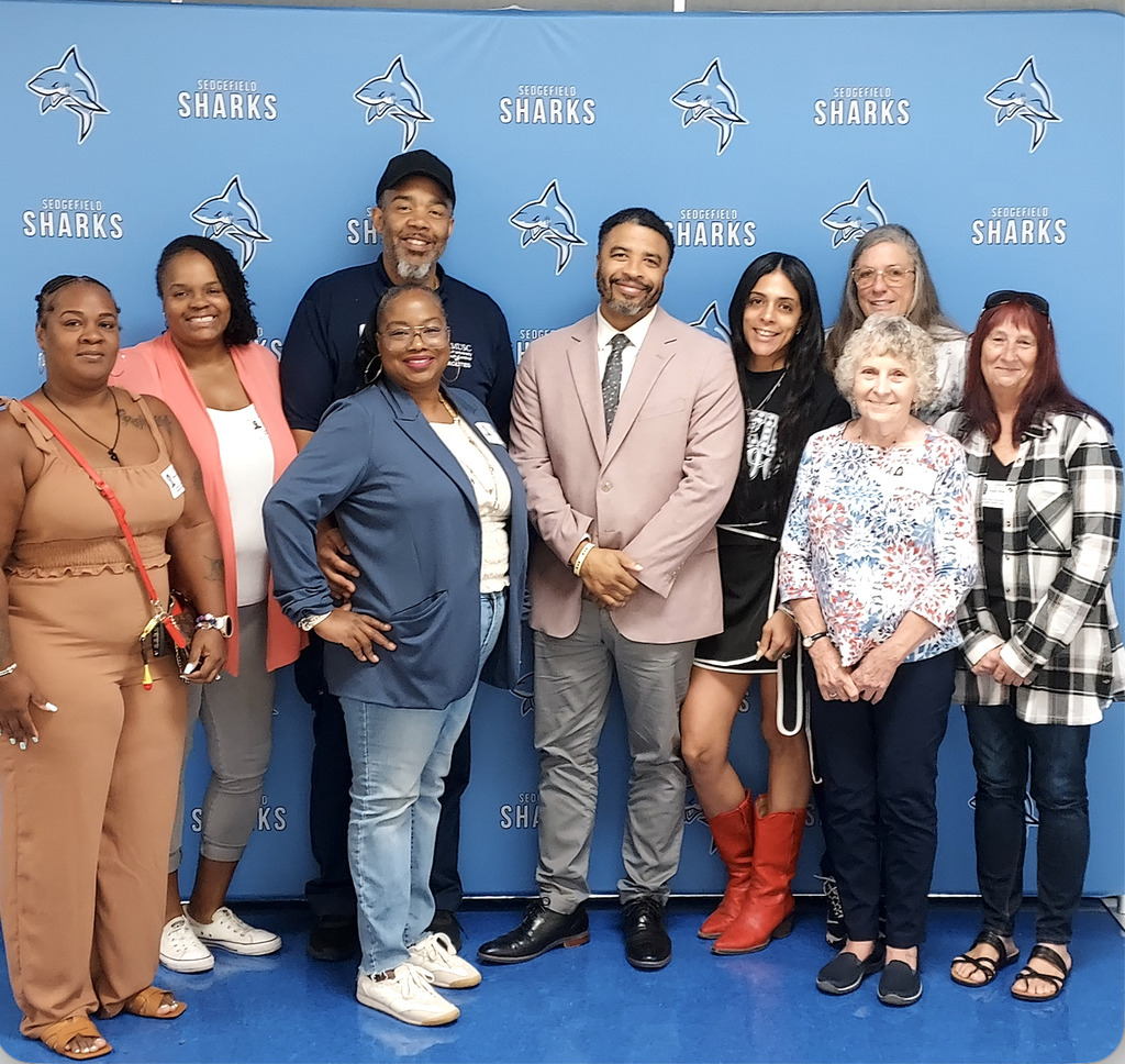 Eleven people stand in front of a blue wall with white Sharks logos. The man in a hat is in the center.