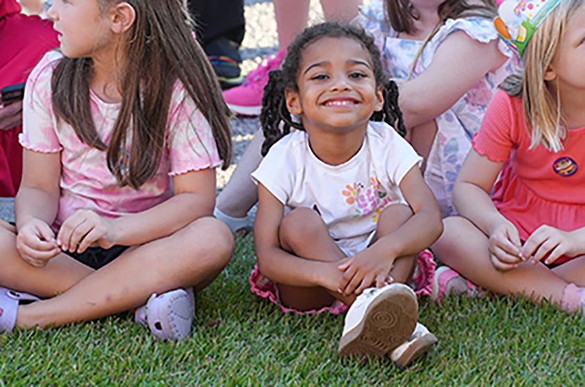 Children sit on grass, smiling. One wears a pink shirt, another a white shirt with flowers, and the third a red dress.