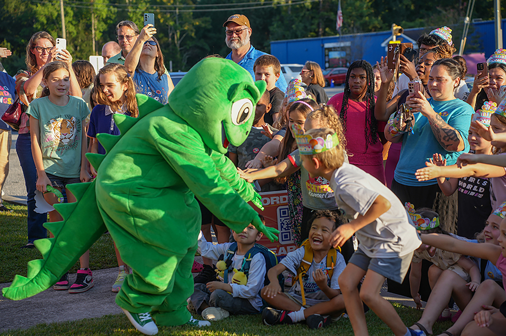 A person in a green dinosaur mascot costume greets children in a grassy outdoor area. Some children wear party hats.