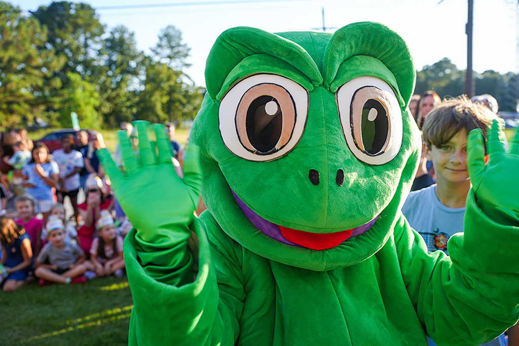 Green frog mascot waving at children seated in a grassy area. Trees and a pole in the background.