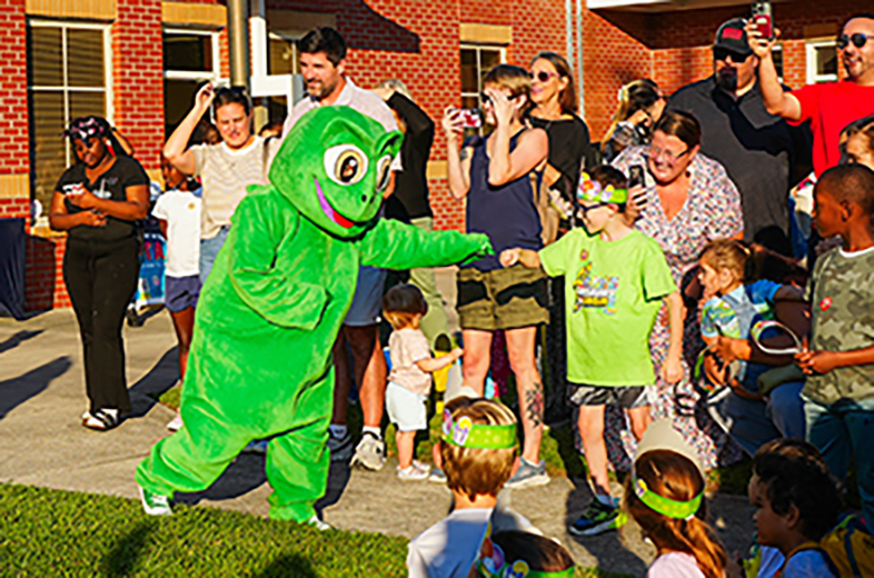 A green mascot entertains children in front of a brick building. Various adults and children stand or sit nearby.