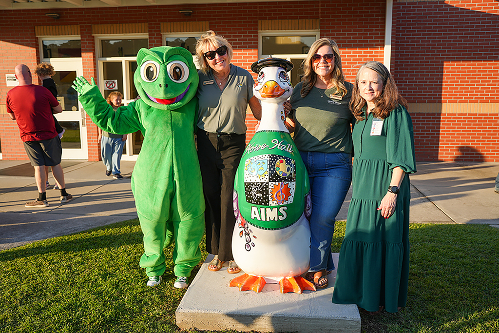 Four individuals pose next to a large statue outside. One person wears a green frog costume and the statue is a large white bird.