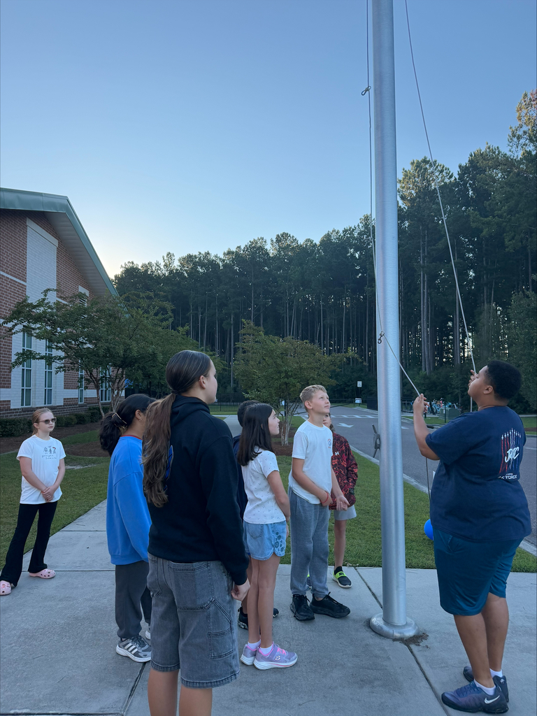 Kids gather in front of a flagpole with one person raising the flag. Trees and a building in the background.