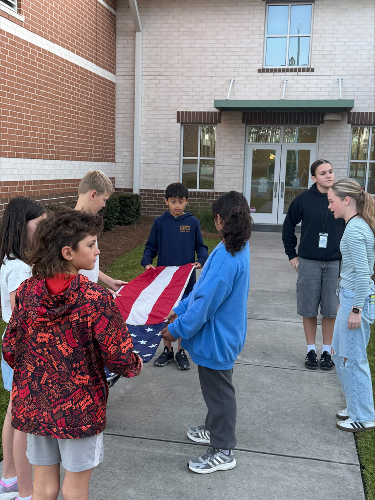 Students stand around an American flag. A brick building with glass windows and doors is in the background.