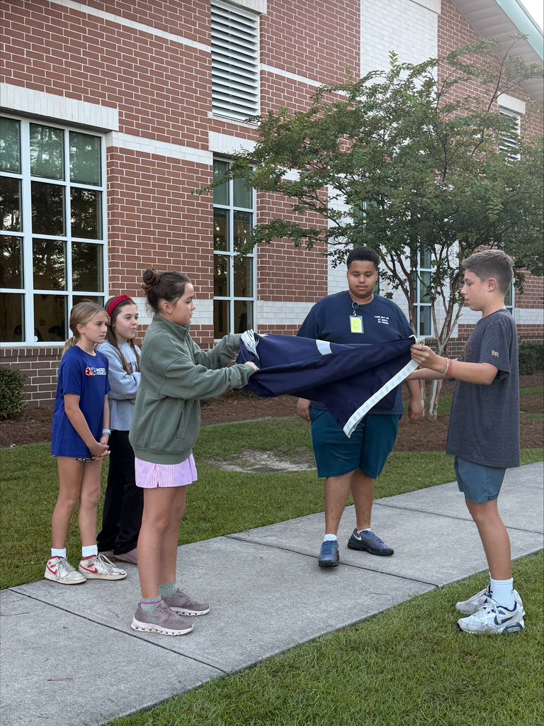 Four children in shorts and sneakers hold up a blue cloth outdoors near a building. Two children hold the ends.