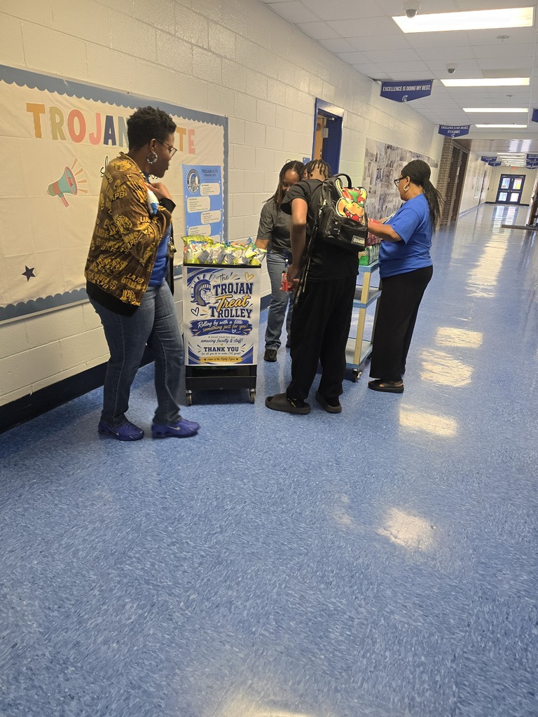 People stand in a hallway, some looking at a cart with food, near a wall with "TROJA" sign.