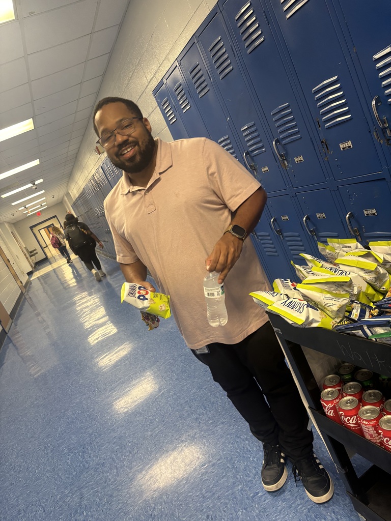 Man in hallway of school with blue lockers, holding snacks and a water bottle, and people walking.