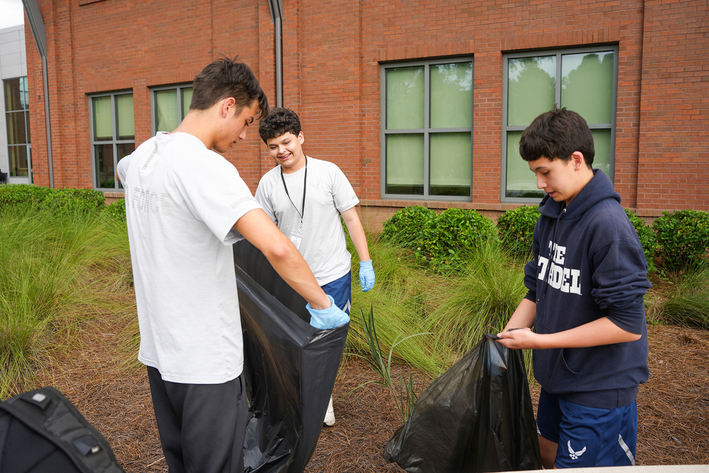 Three individuals with gloves on, standing outside a brick building. One holds a black bag, another looks at it, and the third speaks.