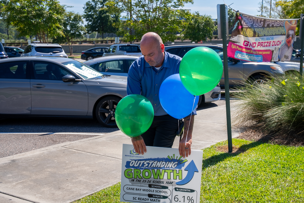 A man holding three balloons stands near a sign with "OUTSTANDING GROWTH" and "6.1%" on a grassy lawn.
