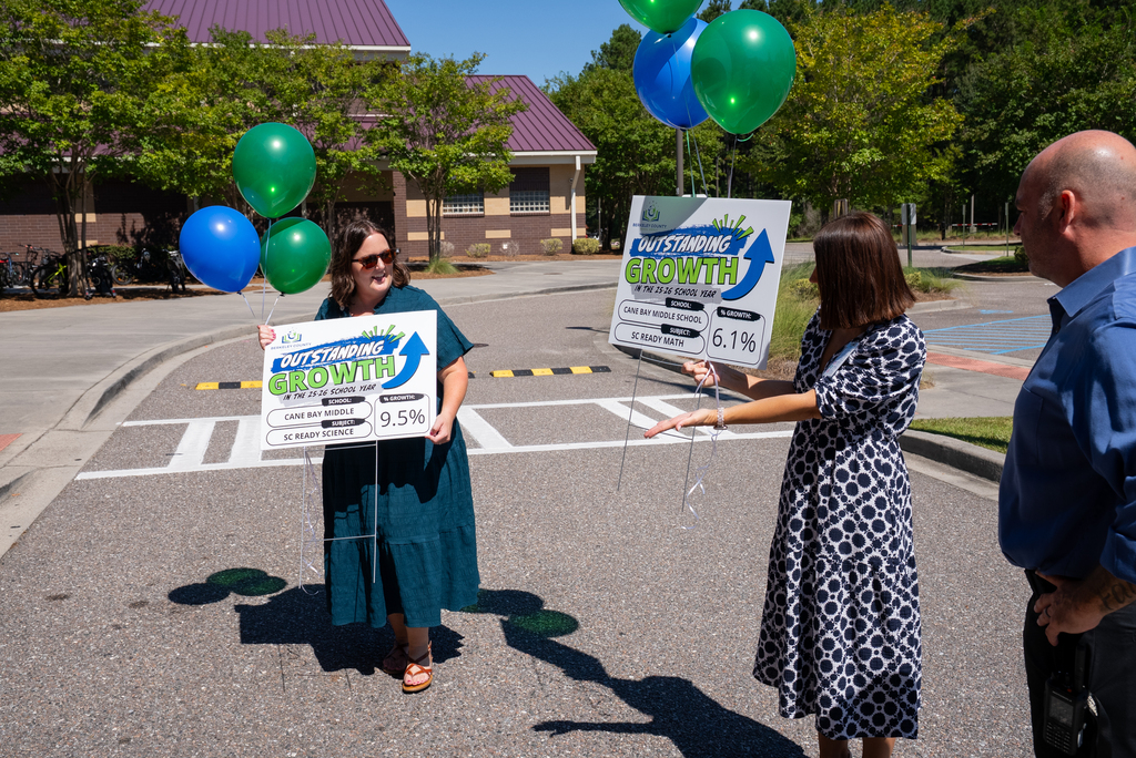 Three adults, each holding a sign, stand in a street. Behind them, a building has trees and balloons.