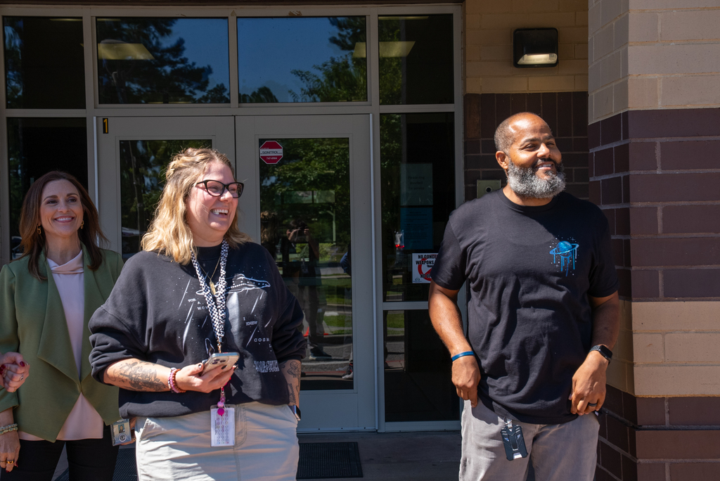 Three people outside a building. A woman with blonde hair and glasses, a man with a beard, and a woman with a green top.