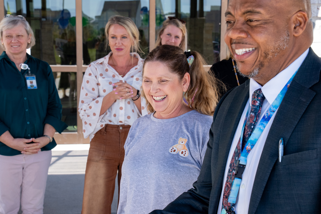 Several people in business attire stand outside a building. A woman in a gray shirt smiles as a man next to her shakes her hand.