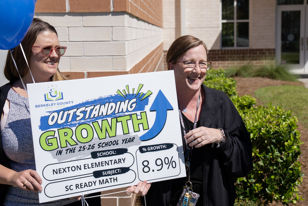 Two women holding a sign that says "Outstanding Growth" in front of a building with greenery.