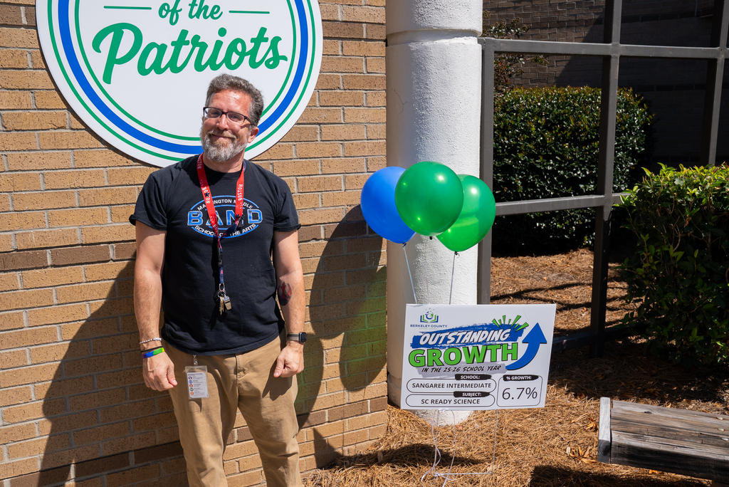A man stands next to a building with a Patriots sign. He wears a black shirt, khaki pants, and a beard.
