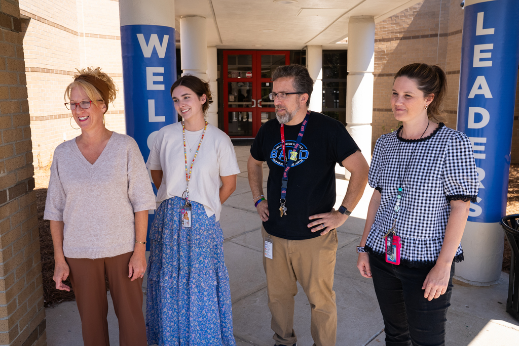 Four adults stand together in front of a building. Three women wear glasses. A blue sign behind them reads "WELL LEADERS."
