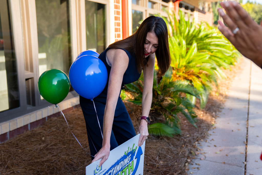 A woman in a blue dress holds a sign with balloons, bending over by a building.