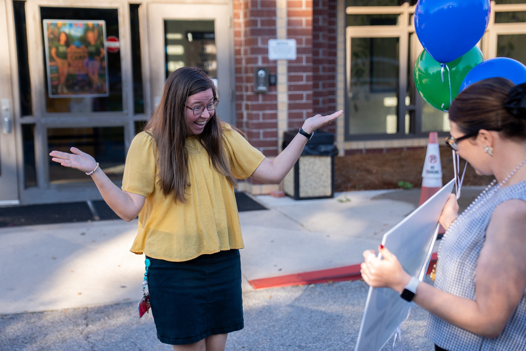 Two women stand outside a brick building, one gesturing while the other holds a clipboard. A traffic cone and balloons are nearby.