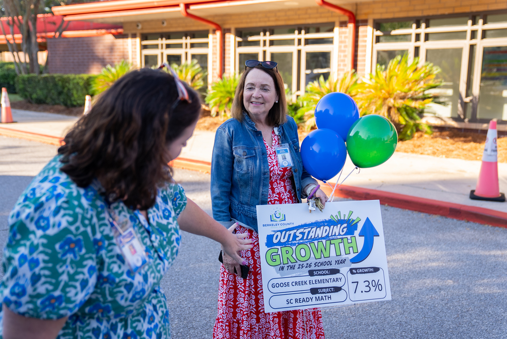 Two women hold a sign for outstanding growth in front of a building. One woman smiles, holding balloons.