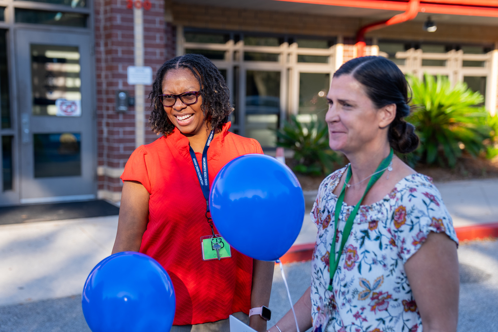 Two women outside a building, one holding two blue balloons, both smiling. Behind them is a brick wall and greenery.