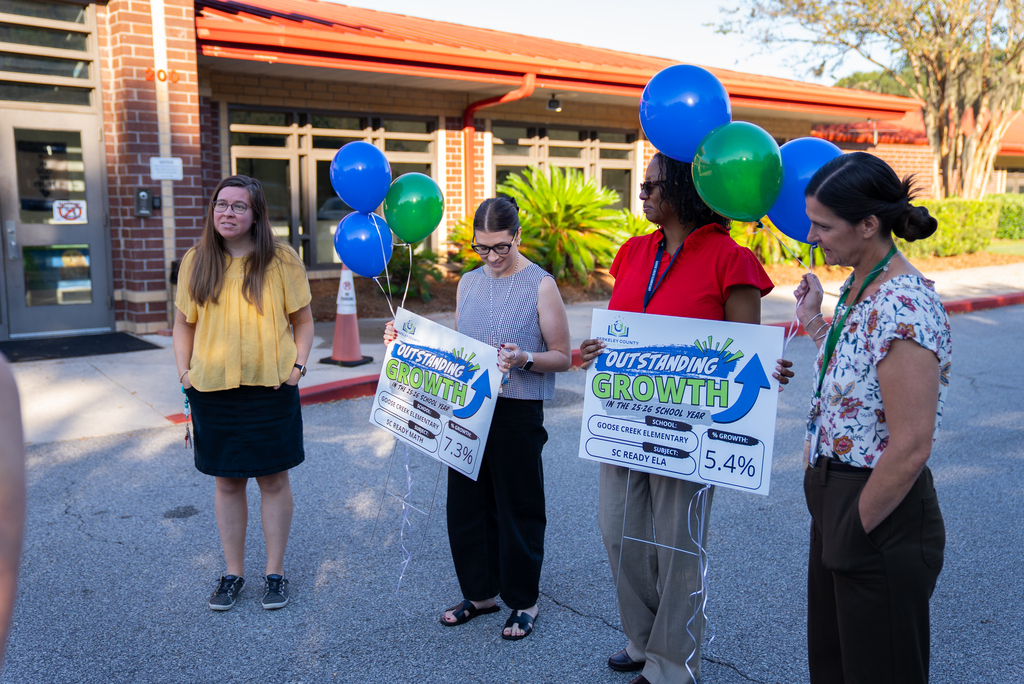Four women stand with signs in front of a brick building. The signs read "Outstanding Growth" with one sign showing 5.4%.