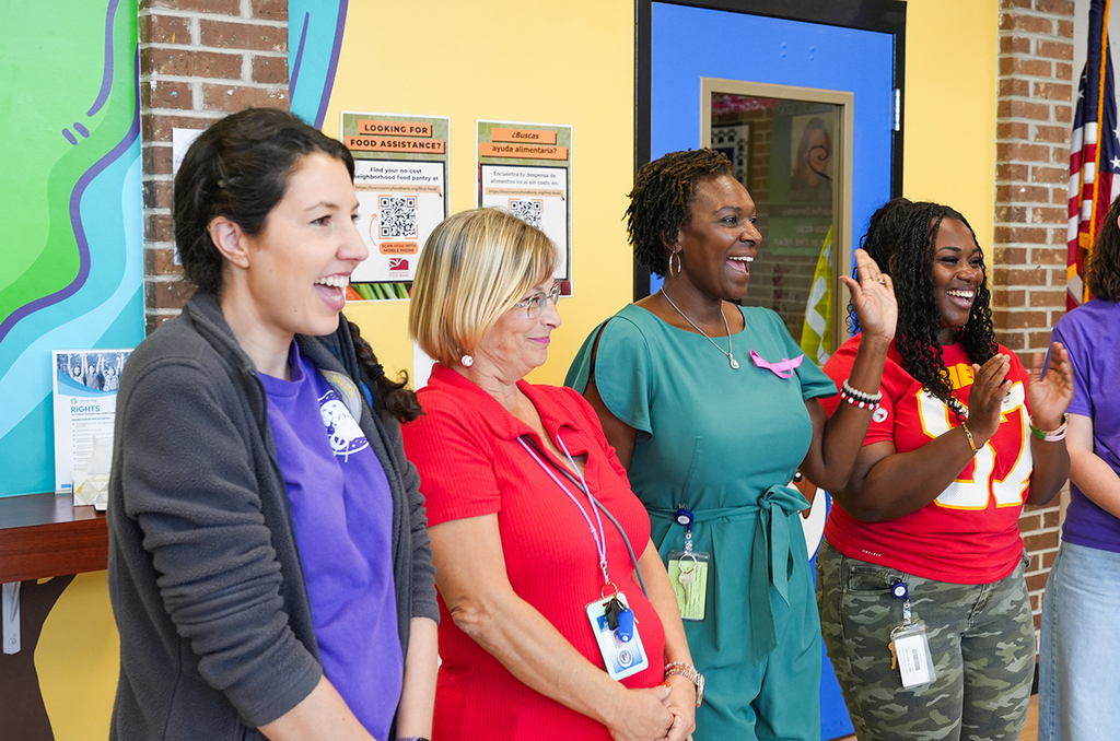 Four women in a colorful room with a brick wall. One waves. One wears a green dress, one a red shirt.