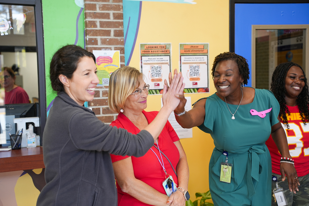 Four women stand together in a colorfully decorated room. Two women give each other high fives.