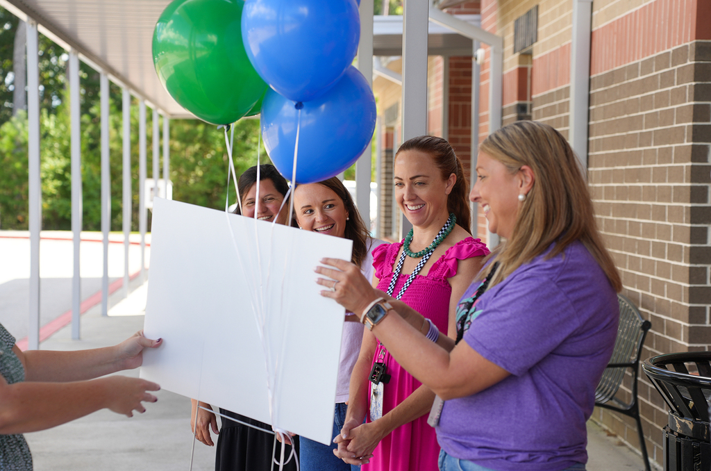 A group of four women holding a large white sign and balloons. A brick wall and trees are in the background.