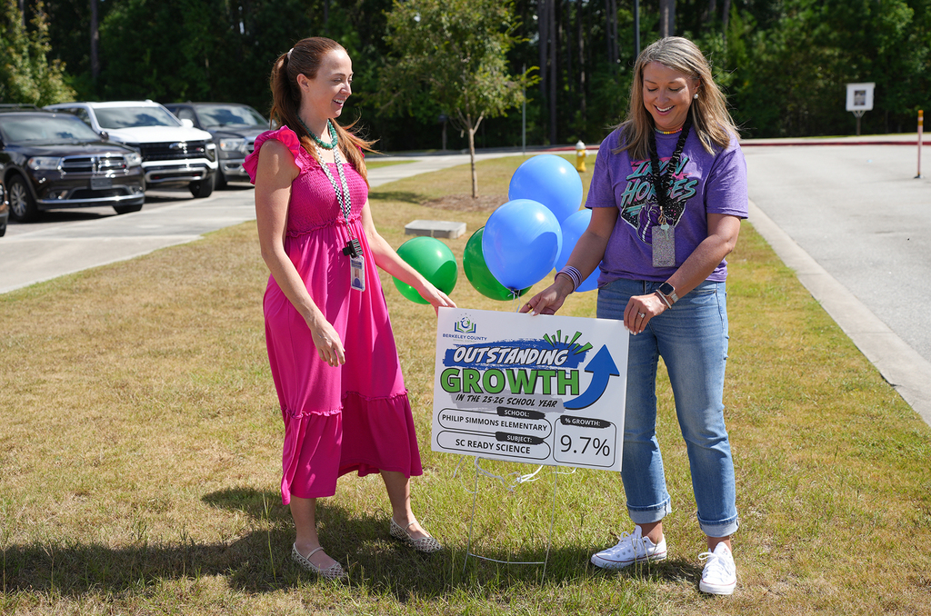 Two women hold a sign with "Outstanding Growth 9.7%" and colorful balloons. They are in a grassy area with parked cars.