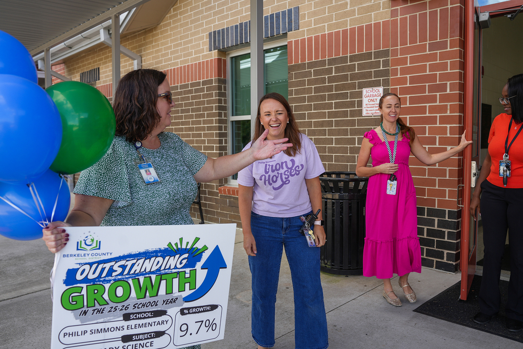 Four women stand outside a brick building with one holding a sign that reads "Outstanding Growth" with balloons.