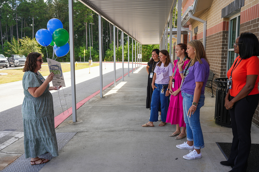 A group of people stand on a school porch. One holds a board, and others watch. There are balloons behind them.